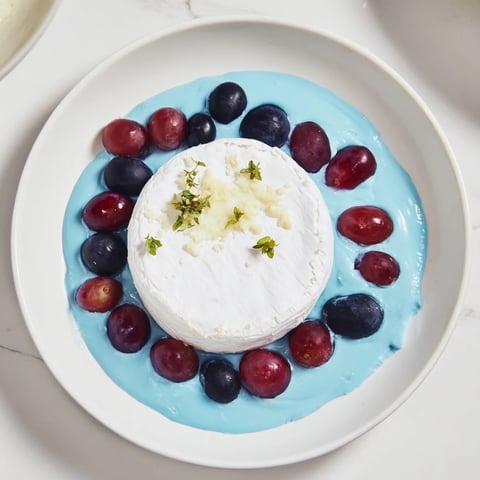 A creamy "Floating Island" cheese wheel sits atop blue yogurt, surrounded by dark grapes.